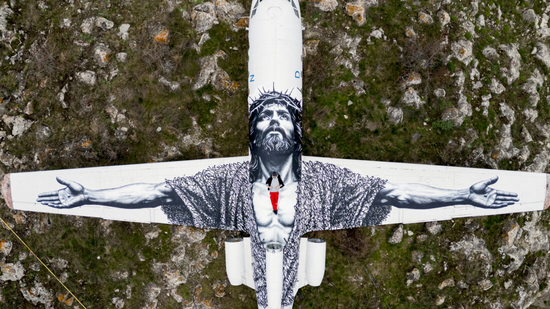 Der Berner Künstler Vierwind alias Micha Haeni in roter Hose auf seinem Kunstwerk: der gekreuzigte Christus, gemalt auf auf ein Flugzeug und betrachtet aus der Himmelsperspektive. Für Haeni symbolisiert es, dass an Himmelfahrt Gott herabsteigt zu den Menschen. 20260514152057863.jpg